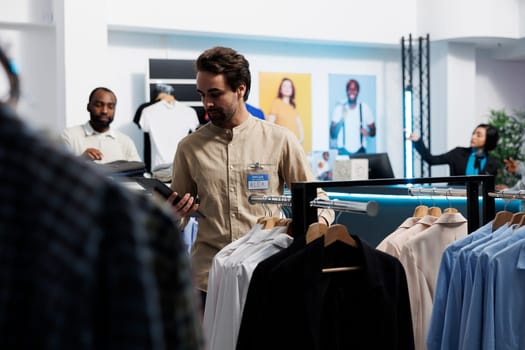 Clothing store manager using digital tablet and organizing merchandise to attract customers. Shopping center employee managing inventory and examining apparel hanging on rack