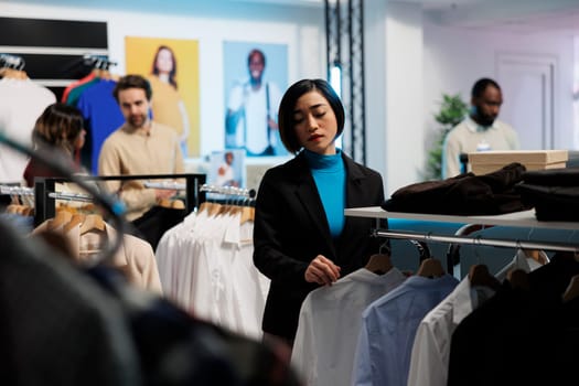 Young asian woman exploring rack with trendy apparel while choosing modern fashionable outfit in shopping mall. Clothing store customer examining formal shirts style in showroom