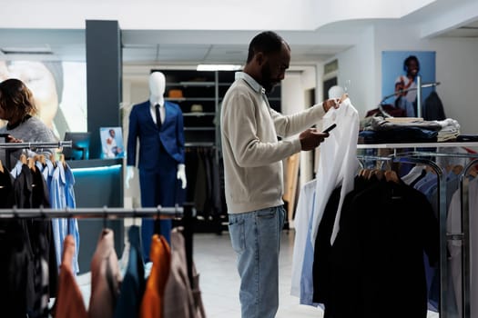 Retail market boutique customer examining hanging garment on rack while selecting size and style in smartphone app. Man choosing apparel in clothing store and checking discount on mobile website