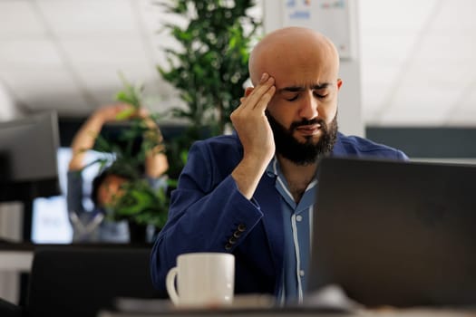 Tired arab businessman with headache working in business office. Overworked executive manager suffering from migraine and rubbing temples while analyzing start up company report on laptop