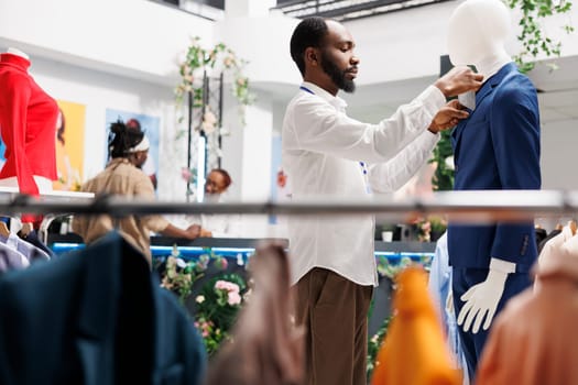 Clothing store employee adjusting male suit on mannequin while working in shopping mall. Fashion boutique african american assistant fastening formal jacket on dummy model