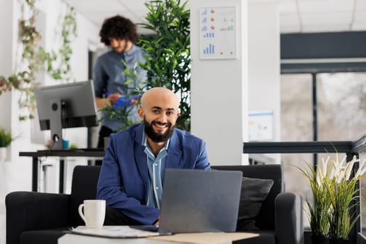 Smiling business entrepreneur working on laptop and looking at camera in start up company office. Successful project manager using portable computer while sitting on couch portrait