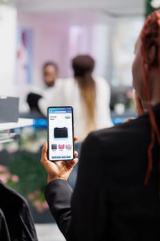 Female client browsing clothing app in fashion boutique, looking at modern clothes and merchandise on online website. Woman using smartphone to buy items on electronic digital store.