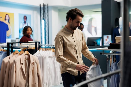 Man selecting fashionable outfit in clothing store and browsing rack with shirts. Young customer examining apparel on hangers while choosing formal wear in shopping mall department