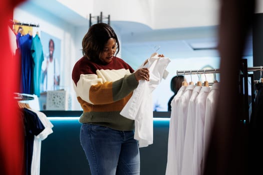 Fashion boutique client holding white shirt on hanger and checking fabric quality. African american woman examining apparel rack while searching for formal outfit in clothing store