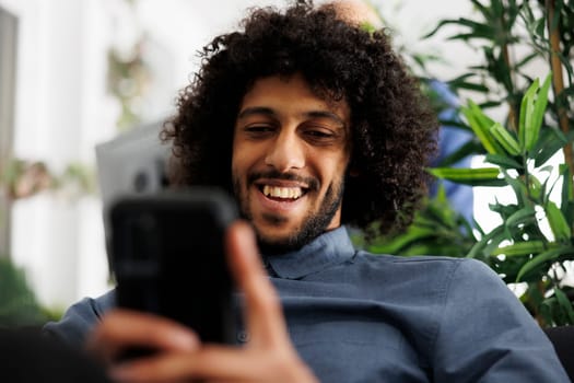 Arab businessman browsing internet on smartphone while relaxing in start up business office. Smiling young corporate employee scrolling social media on mobile phone at work