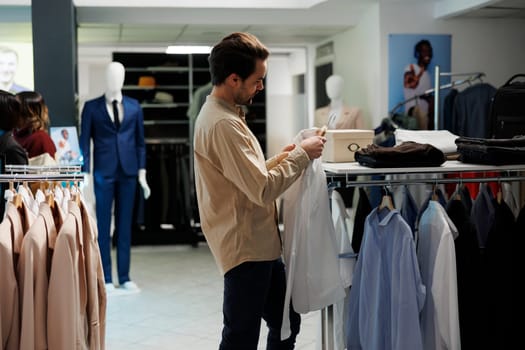 Clothing store customer holding formal shirt on hanger and looking at size label. Young stylish man checking apparel and choosing fashionable garment in shopping mall department