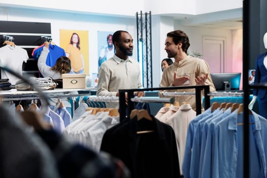 Clothing store young man worker showcasing trendy stylish apparel on rack to client. Retail market fashion boutique african american customer holding outfit hanger and talking with consultant