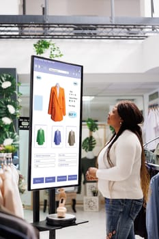 Shopper choosing clothes on display, using touch screen kiosk monitor in trendy mall boutique. Young store client shopping for clothing line with self ordering interactive board service.