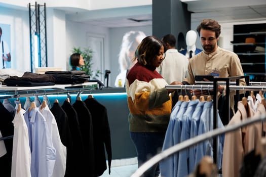 Woman getting help from clothing store employee while choosing casual jacket in boutique. African american shopper asking assistant for advice while shopping in garment showroom