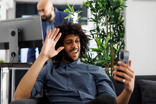 Happy arab businessman waving hi while talking on smartphone online meeting in start up business office. Company executive manager greeting remote coworker on mobile phone videocall