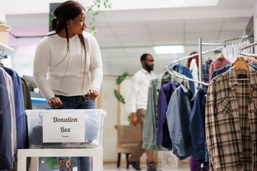 African american woman choosing clothes to put in donation box in shopping center. Fashion boutique client donating new and used apprel in plastic container for humanitarian aid