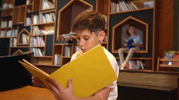 Caucasian boy reading a book while group of smart students sitting at library. Attractive child studying, learning from novel or textbook while children talking, chitchat about education. Erudition.