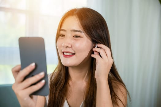 Happy relaxed female touching earbuds listen song on mobile phone in bright living room at home, Lifestyle Asian young woman wearing wireless earphones listening to favorite song on her smartphone