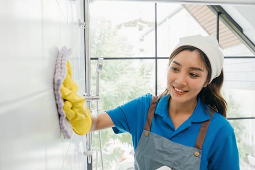 Maid wearing protective gloves wipes dust off ceramic tiles with a microfiber cloth in the bathroom. Her routine cleaning emphasizes purity and hygiene in commercial cleaning.