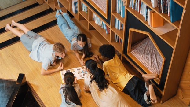 Group of diverse children lying down in circle while reading a book at library. Top view of girl sharing a magazine while pointing at interested topic and talking with lovely friends. Edification.