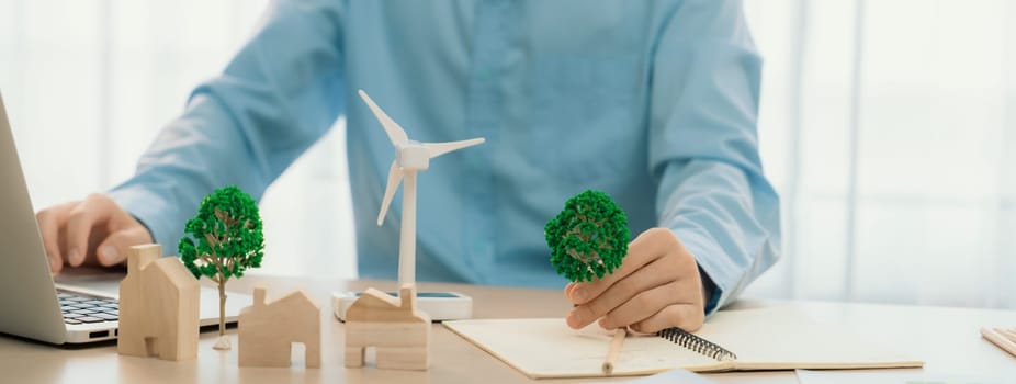 Professional architect plans to use green design in an eco house. Skilled engineer design an eco house blueprint by using laptop on table with house and windmill model scatter around. Delineation.