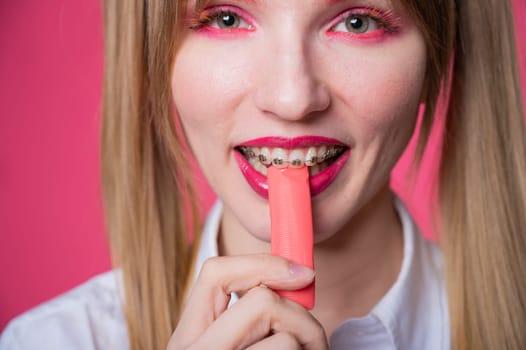 Portrait of a young woman with braces and bright makeup chewing gum on a pink background