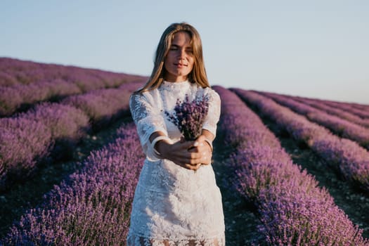 Close up portrait of young beautiful woman in a white dress and a hat is walking in the lavender field and smelling lavender bouquet.