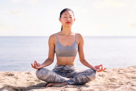 young asian woman doing meditation at beach sitting with legs crossed, concept of mental relaxation and healthy lifestyle