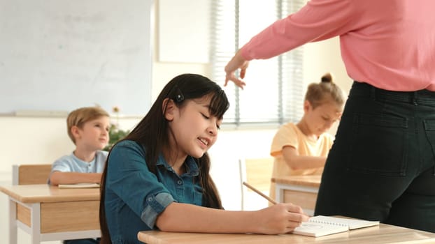 Happy Attractive student writing plan or taking a note during class at elementary class while teacher choosing student to write answer on blackboard. Diverse children attend in classroom. Pedagogy.