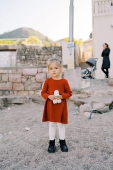 Little girl with a soft toy in her hands stands near a stone fence on the beach. High quality photo
