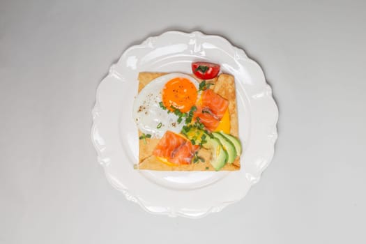 Close-up shot of a crepe with smoked salmon, avocado, egg, and tomatoes on a white plate. Square shape, elegant presentation.