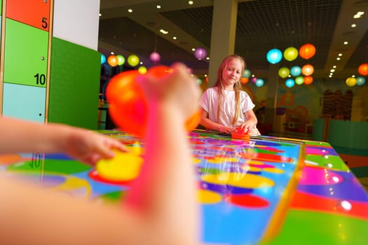 A joyful young girl is captured engaging in a playful board game, surrounded by bright and whimsical decorations in a colorful playroom setting. A hand is seen reaching for a game piece on the reflective, rainbow-hued surface, emphasizing the dynamic and interactive aspects of the game.