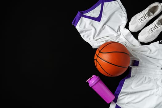 A collection of basketball essentials is neatly arranged against a stark, dark background, including a vibrant orange basketball, white sneakers with purple detailing, a white jersey with purple accents, and a purple water bottle, suggesting readiness for an athletic training session or a game.