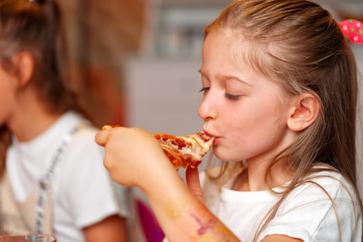 A young girl with light hair is captured in the moment of eating a slice of cheesy pizza, while another child is seated next to her, slightly out of focus. The scene suggests a casual birthday party setting with children enjoying food.