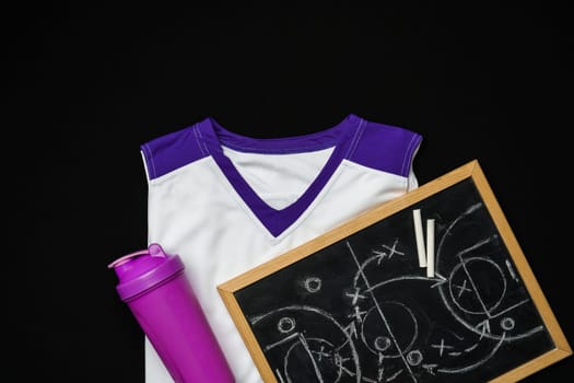 A sports uniform with white and purple colors is laid out beside a purple water bottle and a chalkboard with chalk pieces. The scene suggests preparation for a sporting event or a team strategy session, possibly in a locker room or a team meeting area.