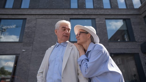 A woman in a hat and glasses hugs an elderly man in a white jacket from behind. Romantic relationships of mature people