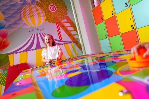 A joyful young girl is captured engaging in a playful board game, surrounded by bright and whimsical decorations in a colorful playroom setting. A hand is seen reaching for a game piece on the reflective, rainbow-hued surface, emphasizing the dynamic and interactive aspects of the game.