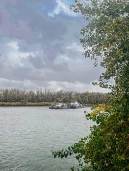 The image captures the serene scene of tugboats navigating the river under a cloudy sky with autumn trees on the shore.