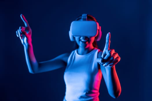 Smart female standing in cyberpunk neon light wear VR headset connecting metaverse, futuristic cyberspace community technology. Elegant woman use finger point generated virtual object. Hallucination.