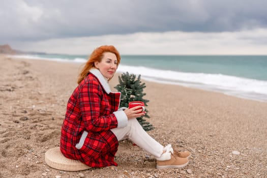 Lady in plaid shirt with a red mug in her hands enjoys beach with Christmas tree. Coastal area. Christmas, New Year holidays concep.