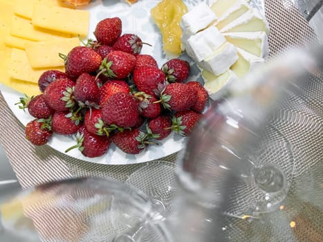 Top view of brie and cheddar cheese slices with fresh strawberries on white dish. Selective focus in a well-lit setting.