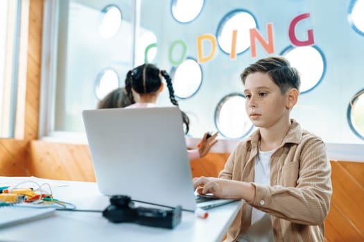 The schoolboy in classroom sitting and learning about engineering remote control with laptop while other child watch outside. Circuit wire, motherboard and notebook put on desk messily. Erudition.