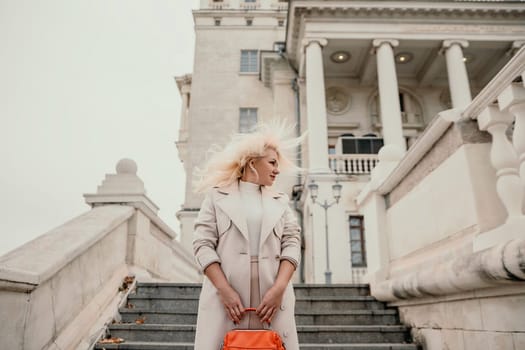 A woman wearing a white coat and blonde hair stands on a set of stairs in front of a large building. She is holding a red purse in her hand. Concept of elegance and sophistication