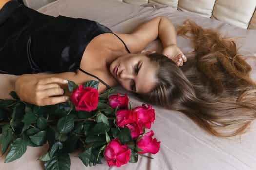 A woman is laying on a bed with a bouquet of red roses next to her. Concept of romance and intimacy, as the woman is surrounded by the flowers and he is in a relaxed and comfortable position