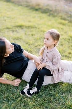 Mom looks at the little girl sitting on her legs lying on a green meadow. High quality photo