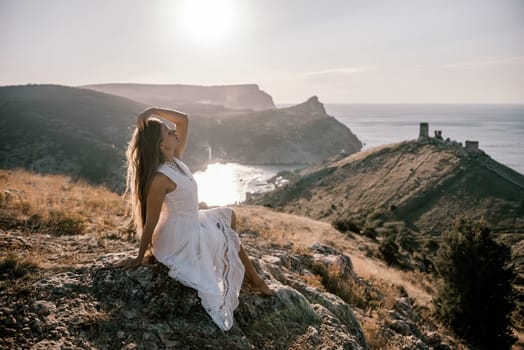 A woman in a white dress is sitting on a rock overlooking a body of water. She is enjoying the view and taking in the scenery