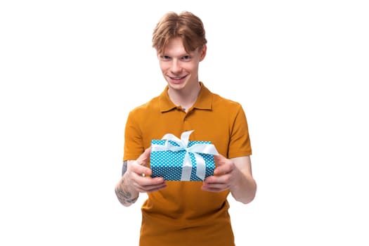 young handsome red-haired guy dressed in an orange t-shirt with a tattoo on his arm holds a gift box for valentine's day.