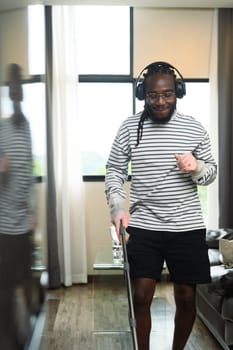 Portrait of happy African man in headphone dancing and singing while cleaning living room.