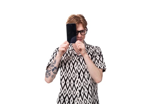 young european male student with red hair dressed in a shirt with a pattern of rhombus shows holds the phone screen forward with a mockup.