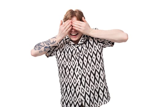 young laughing caucasian ginger man with a tattoo on his arms dressed in a black and white shirt covered his eyes with his hands.