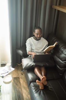 Casual African male freelancer sitting on couch and working online with laptop at home.