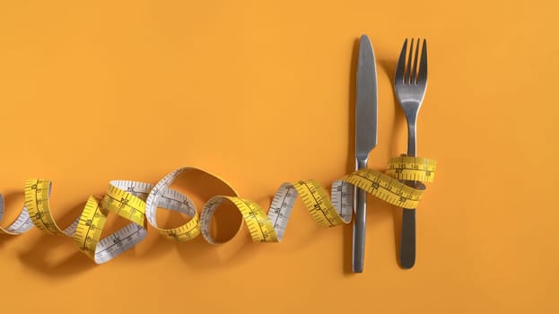 Measuring tape around fork and knife on yellow background. Weight loss and healthy concept.