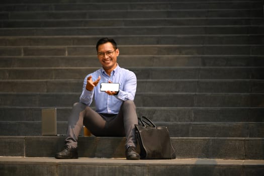 Cheerful businessman sitting on stairs of office building and showing smartphone with blank screen.