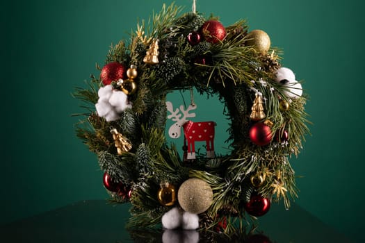 A Christmas wreath with red and green ornaments sits on a wooden table. The wreath is decorated with lights, pine cones, and a red bow, creating a festive display for the holiday season.